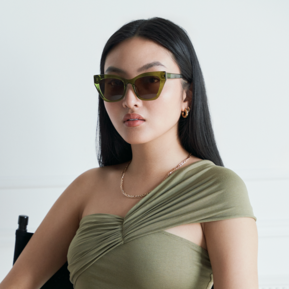 Woman with long black hair wearing KayTran Eyewears Harper - Olive sunglasses, a one-shoulder top, and gold jewelry indoors.