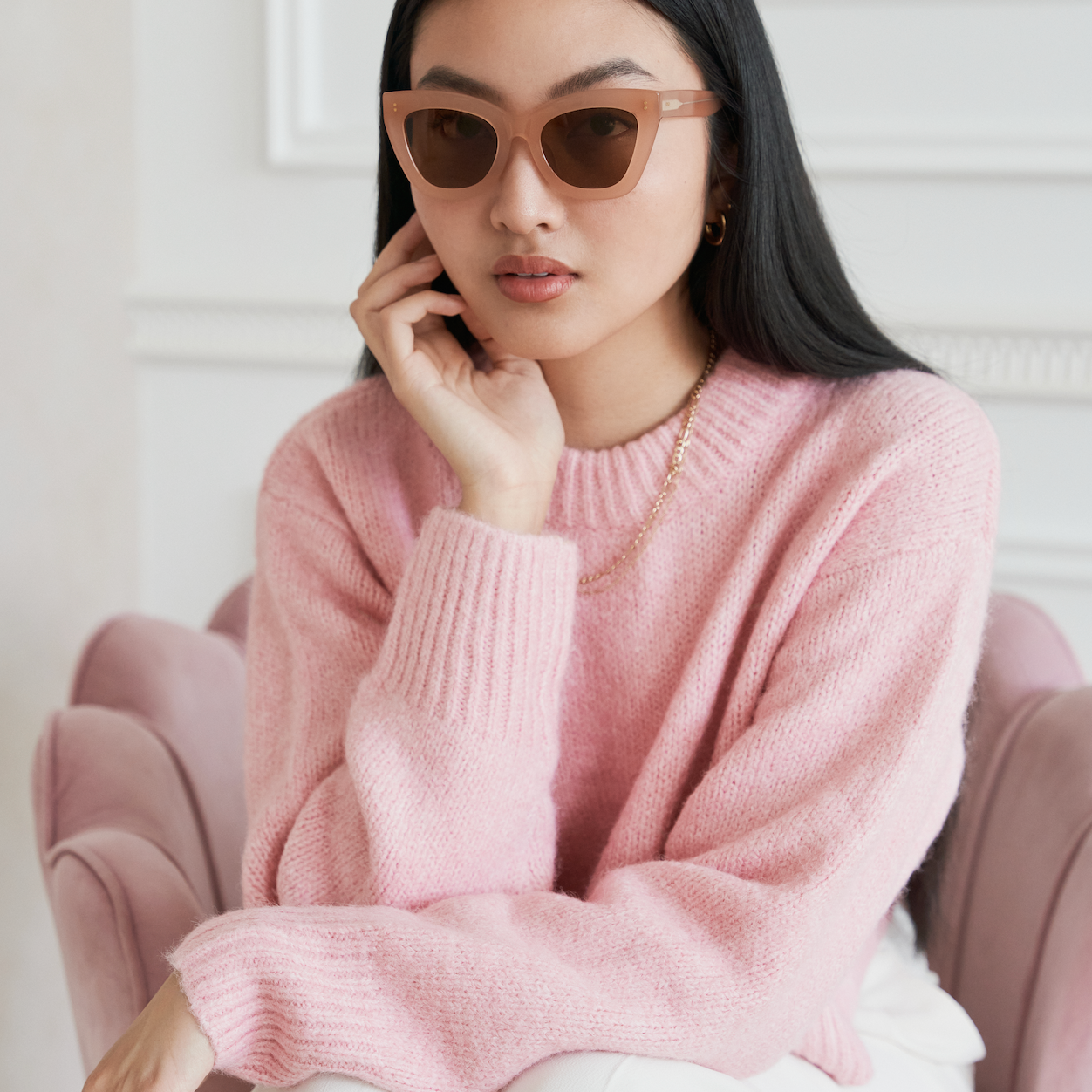 Woman with long black hair wears KayTran Eyewear Arlo - Milky Blush sunglasses and a pink sweater, seated on a pink chair.