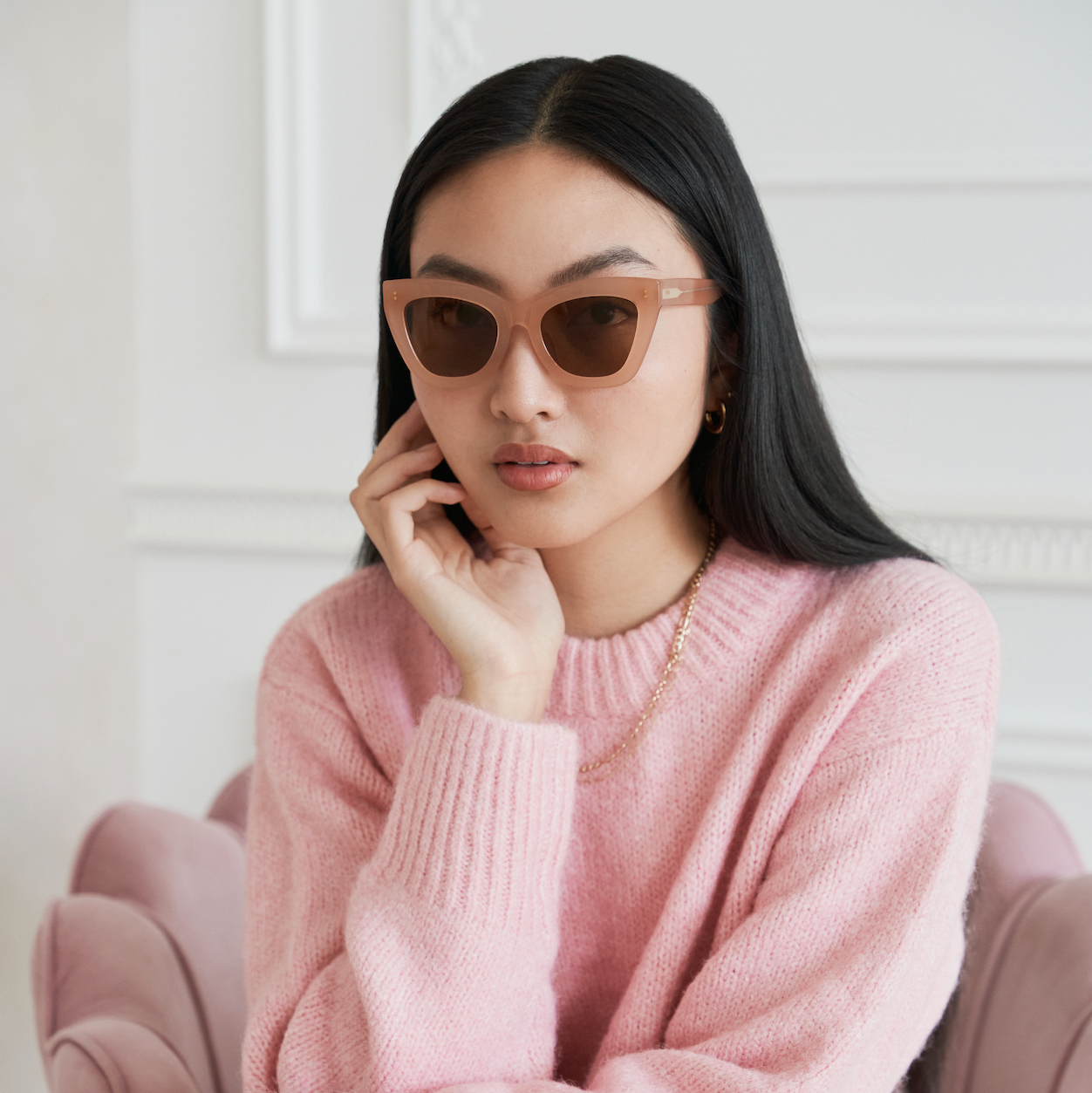 Woman with long black hair wears KayTran Eyewear Arlo - Milky Blush sunglasses and a pink sweater, seated on a pink chair.