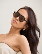 Woman with long brown hair wears KayTran Eyewears Knox - Black & Brown Lens sunglasses against a light background.