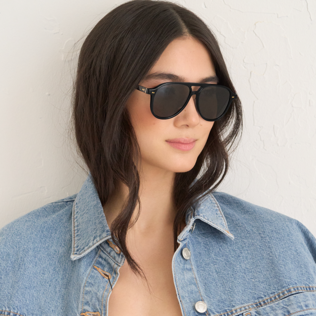 Woman with long dark hair wears KayTran Eyewears Knox - Monochrome sunglasses and a denim jacket against a white wall.