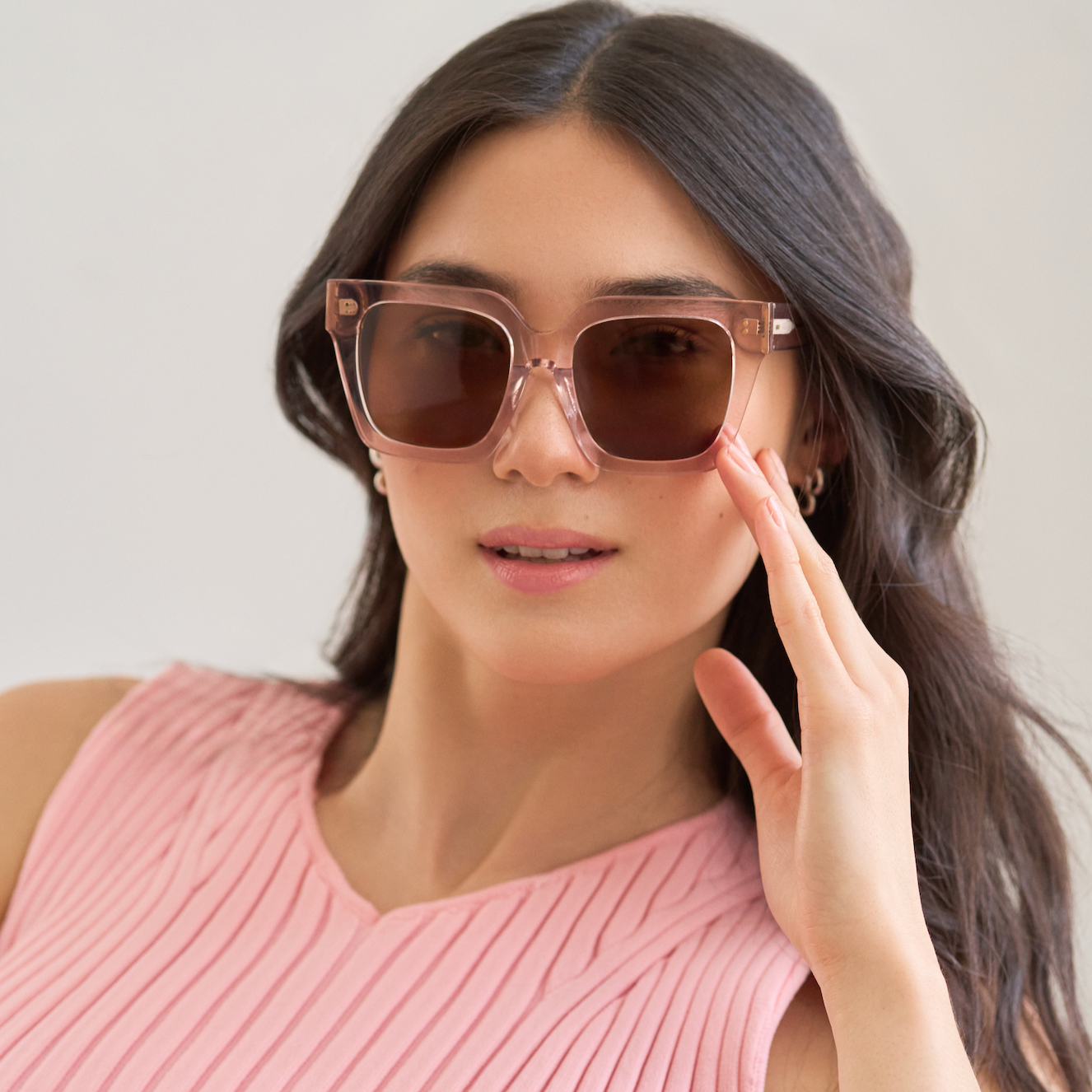 Woman in pink sleeveless top wears KayTran Eyewear Logan - Bloom sunglasses, touching the frame with her right hand.