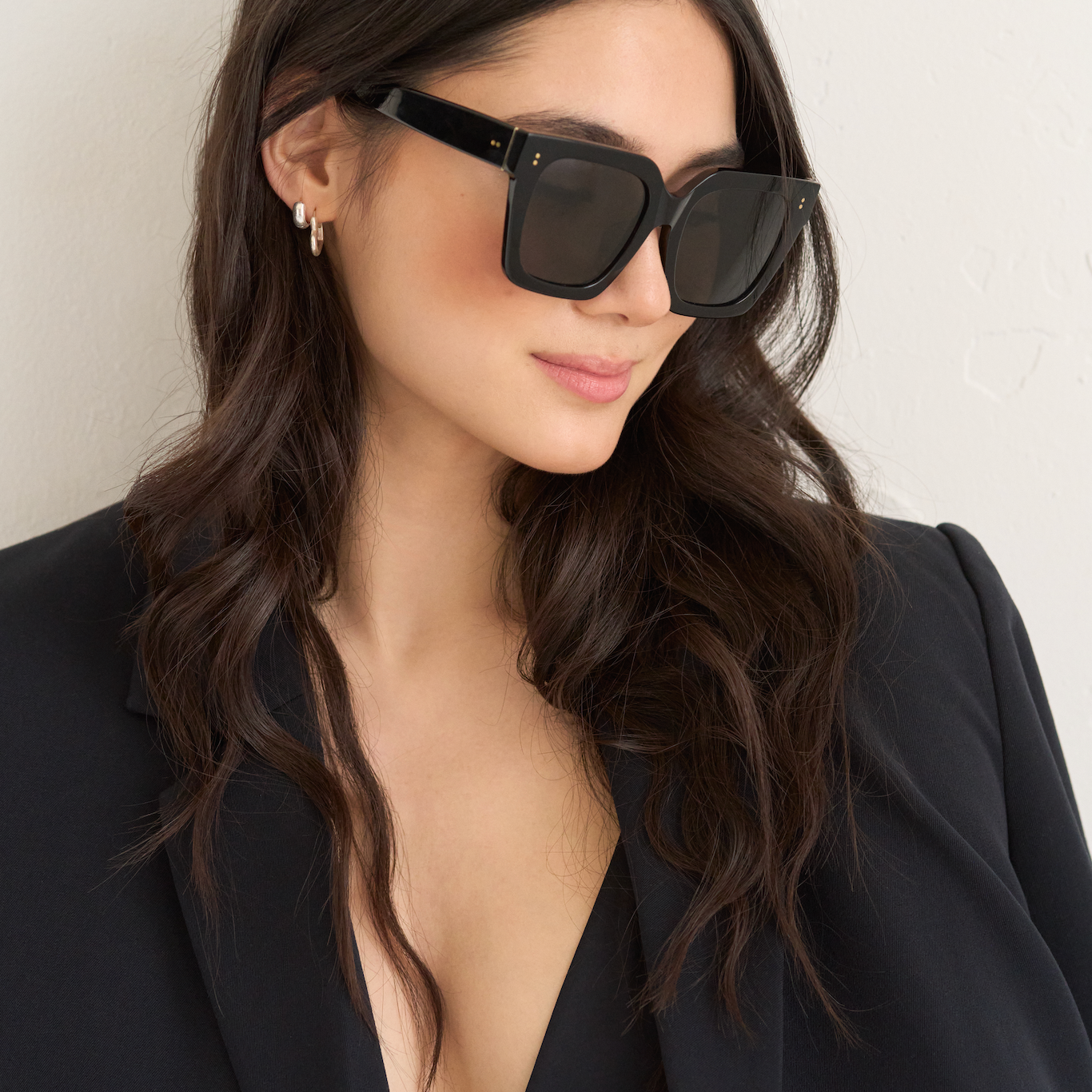 Woman with long wavy hair wears KayTran Eyewear’s Logan - Polished Black sunglasses, black blazer, and top.