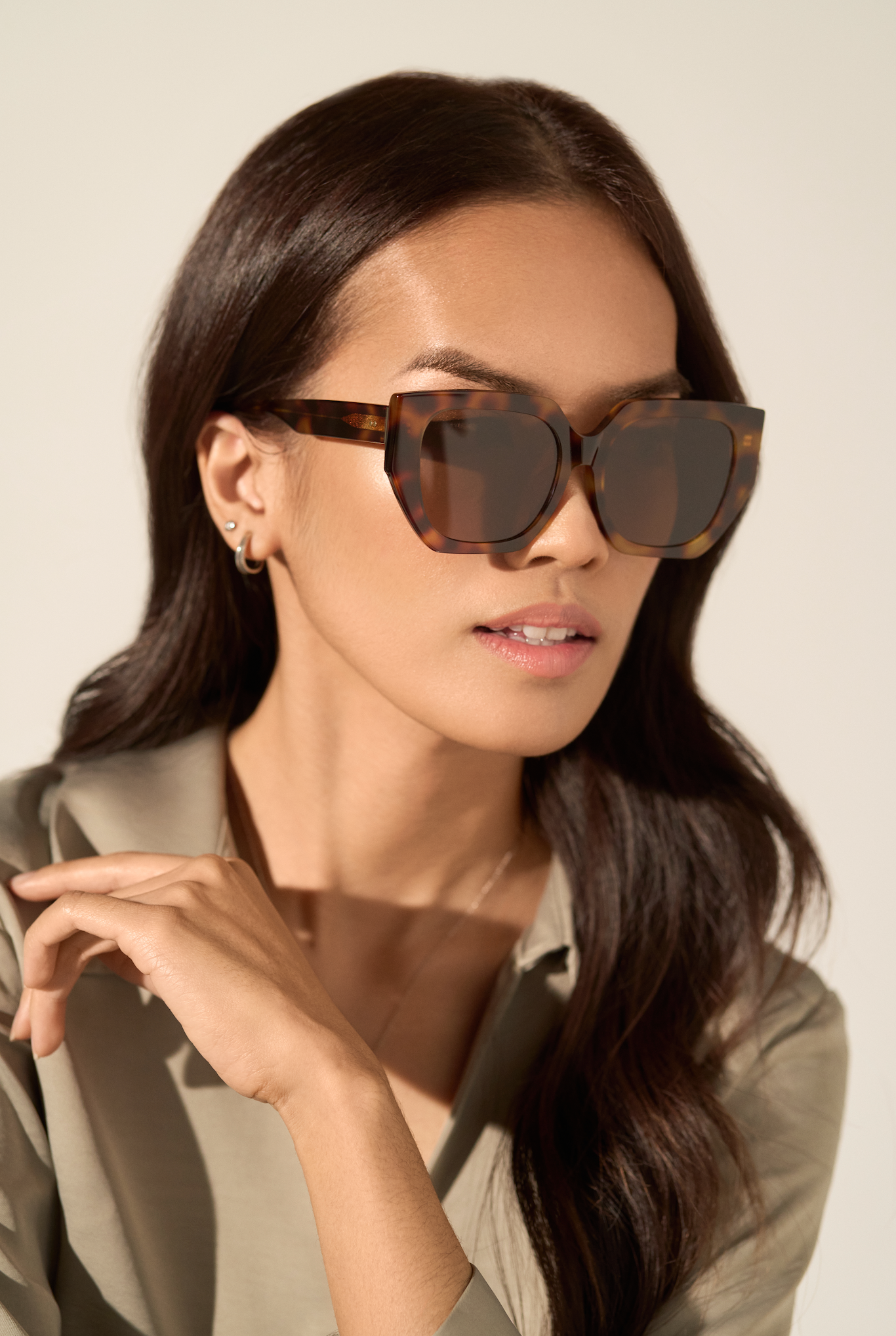 Woman with long dark hair wears KayTran Eyewear’s Luna - Honey Tort sunglasses and a light olive shirt, looking aside.