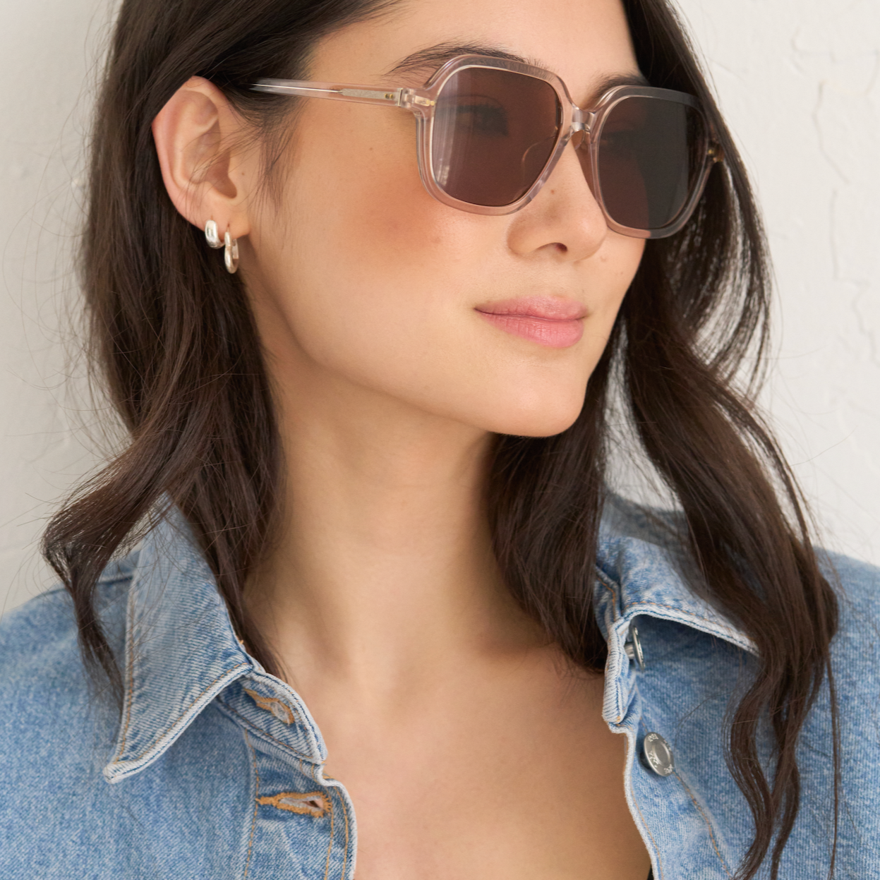 A woman models KayTran Eyewears River - Sand sunglasses with a denim jacket against a light textured wall.