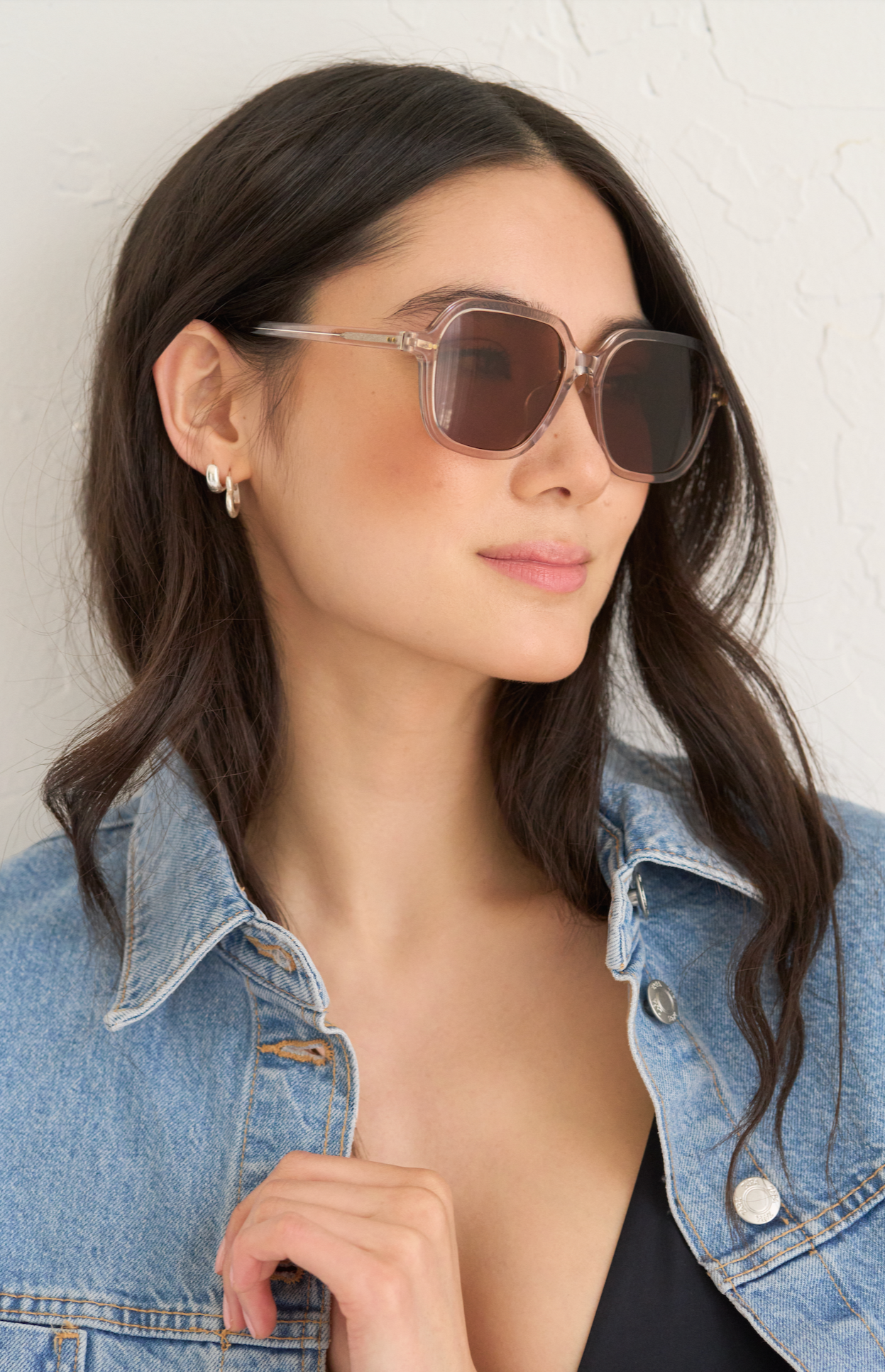 A woman models KayTran Eyewears River - Sand sunglasses with a denim jacket against a light textured wall.