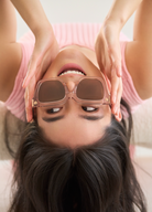 A woman lies upside down, smiling slightly while holding KayTran Eyewears Logan - Bloom square sunglasses over her eyes.