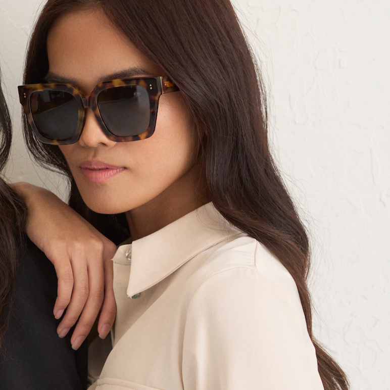 Woman with long brown hair wears KayTran Eyewears Logan - Ember & Grey Lens sunglasses, beige shirt, white wall behind.