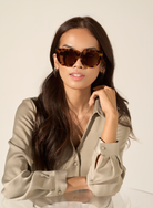Woman with long dark hair wears KayTran Eyewear’s Luna - Honey Tort sunglasses and a beige blouse at a glass table.