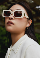Woman wears Brooks - Ecru sunglasses by KayTran Eyewear and a white collared top, standing outdoors.