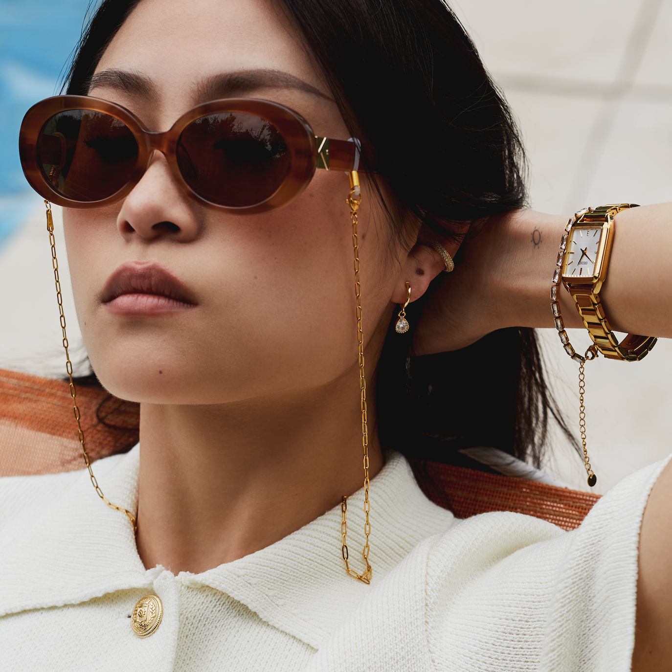 Woman wearing KayTran Eyewear Mara - Latte sunglasses, a white top, gold jewelry, and a gold watch outdoors.