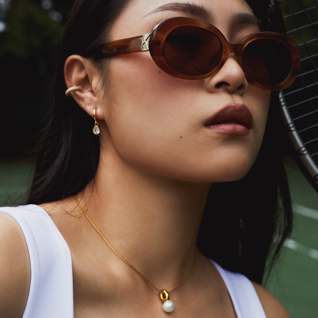 Woman wears KayTran Eyewear Mara - Latte sunglasses, gold jewelry, and a white top on a tennis court with a racket.