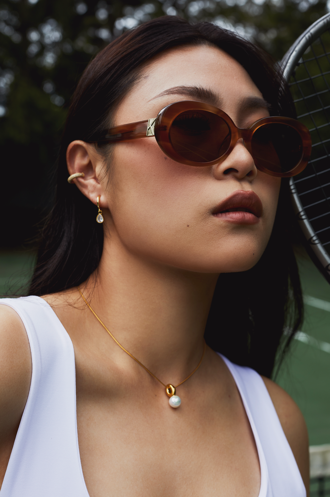 Woman wears KayTran Eyewear Mara - Latte sunglasses, gold jewelry, and a white top on a tennis court with a racket.