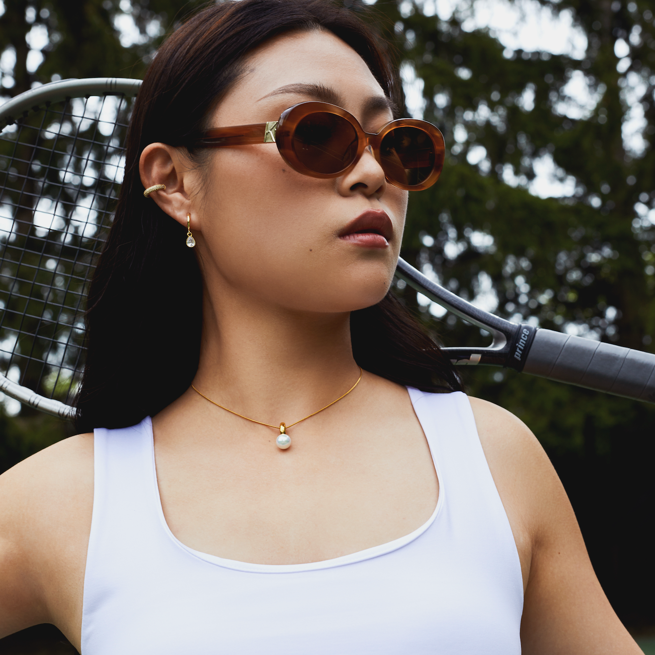 Woman wearing KayTran Eyewear Mara - Latte sunglasses holds a tennis racket on an outdoor court.