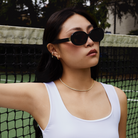 A woman wears KayTran Eyewears Mara - Polished Black sunglasses on an outdoor tennis court.