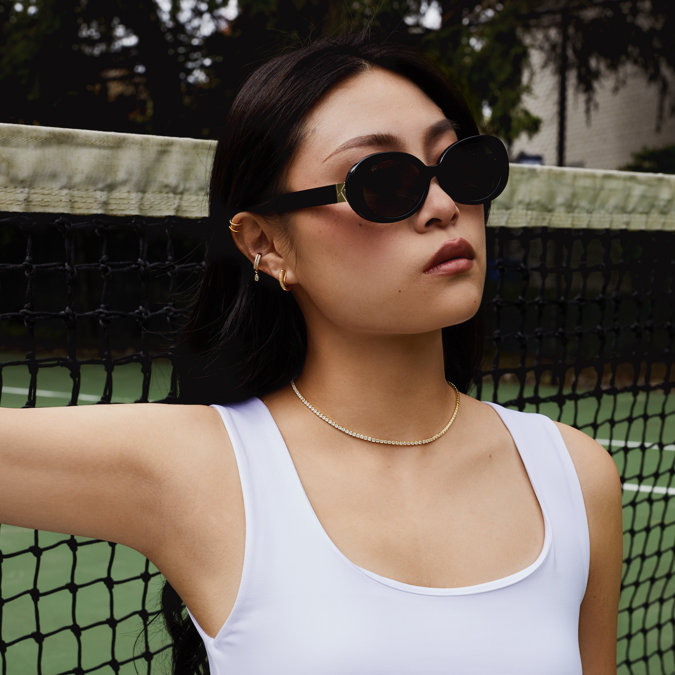 A woman wears KayTran Eyewears Mara - Polished Black sunglasses on an outdoor tennis court.