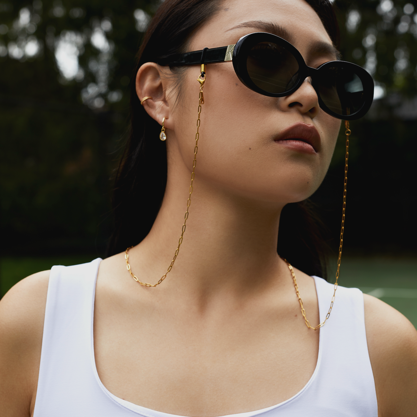 Woman wears Mara - Polished Black sunglasses by KayTran Eyewear, gold chain and earrings, and a white tank top outdoors.