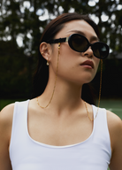 Woman wears Mara - Polished Black sunglasses by KayTran Eyewear, gold chain and earrings, and a white tank top outdoors.