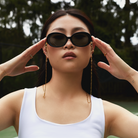 A woman outdoors in a white tank top wears Mara - Polished Black sunglasses by KayTran Eyewear, hands near temples.