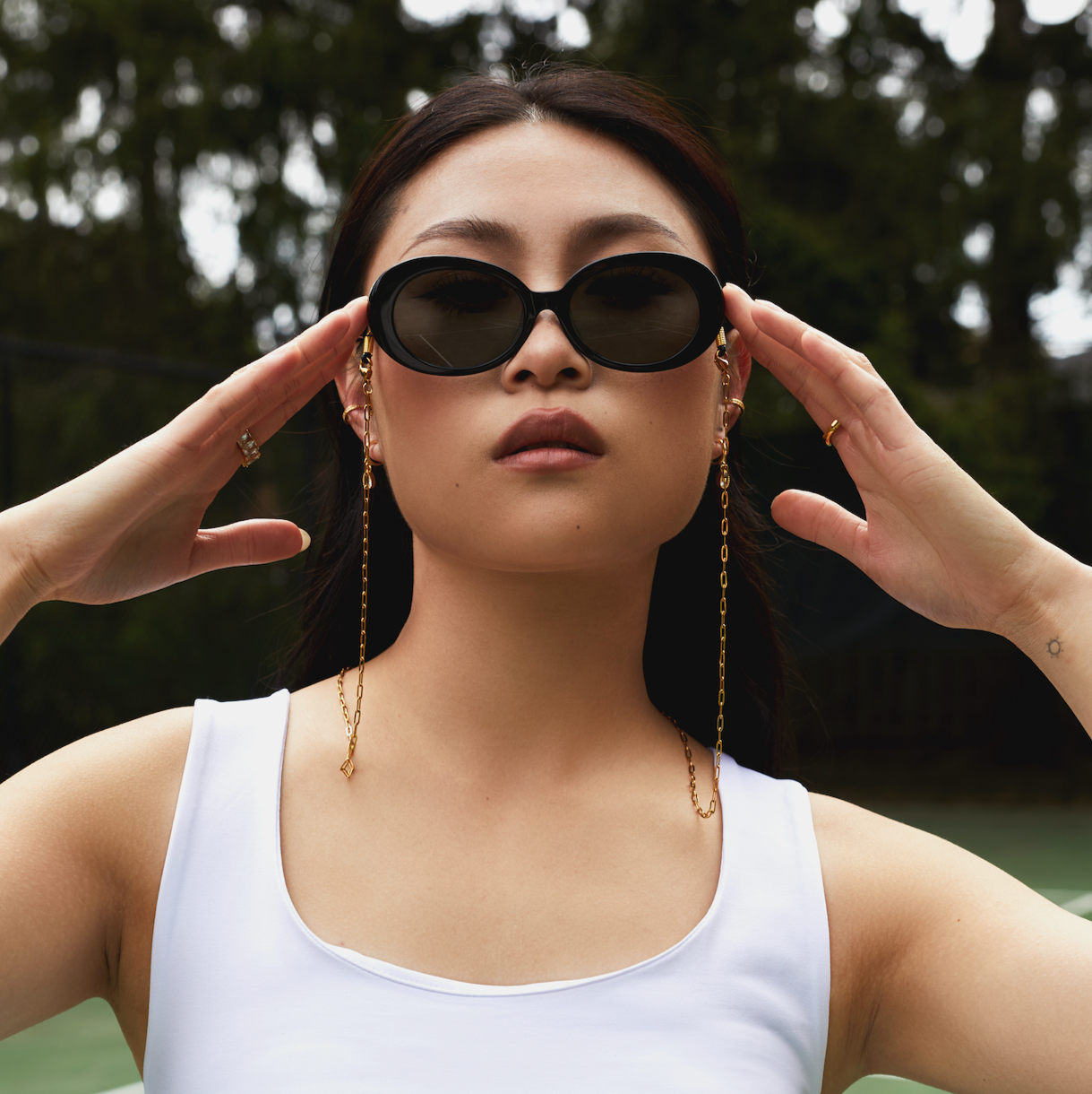 A woman outdoors in a white tank top wears Mara - Polished Black sunglasses by KayTran Eyewear, hands near temples.
