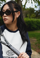 Woman in Mara - Polished Black by KayTran Eyewear, holding a tennis racket and wearing a MANHATTAN, NEW YORK sweatshirt.