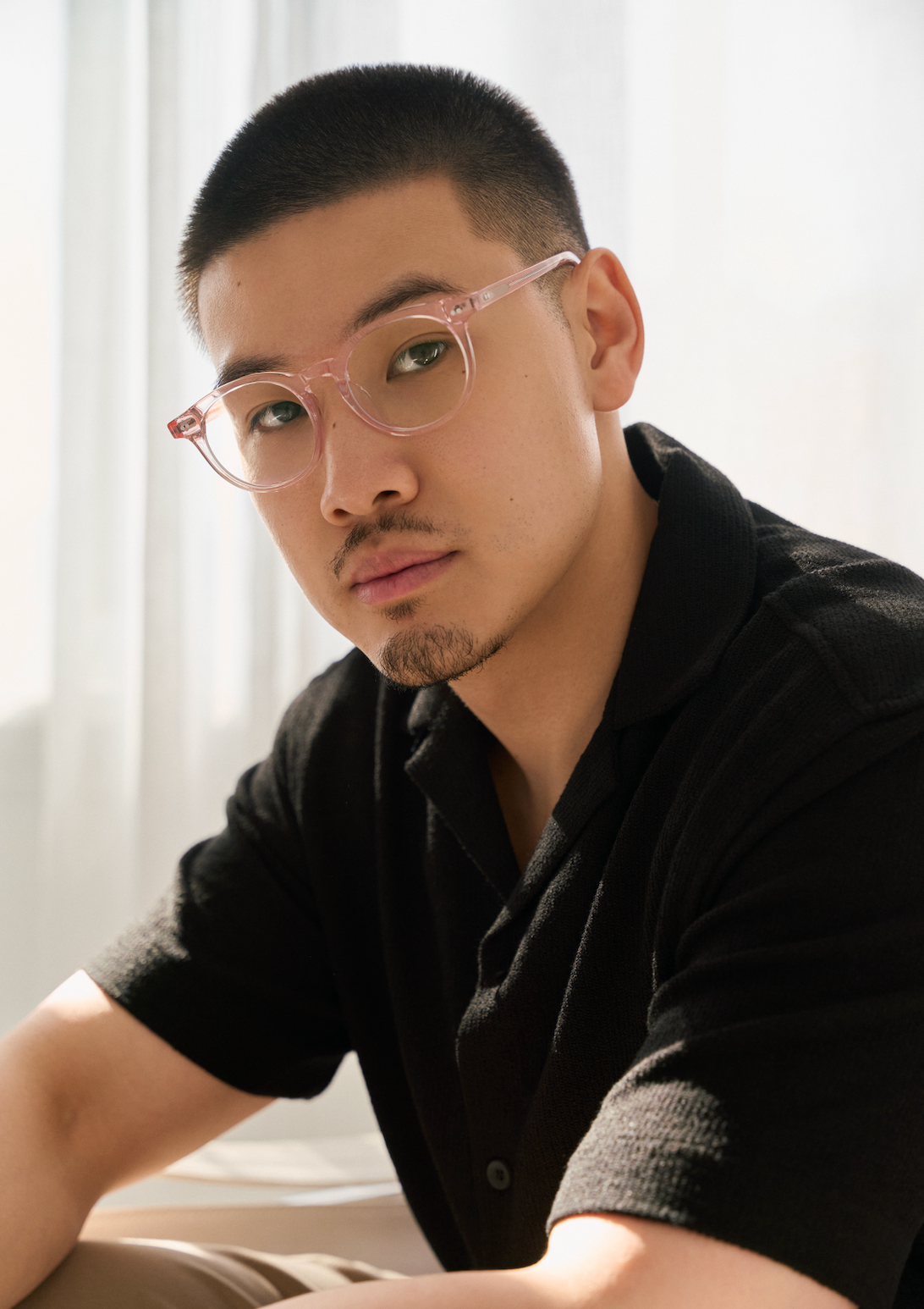 A man with short hair and a goatee wears KayTran Eyewears Bailey - Pastel Pink glasses, sitting by a window indoors.