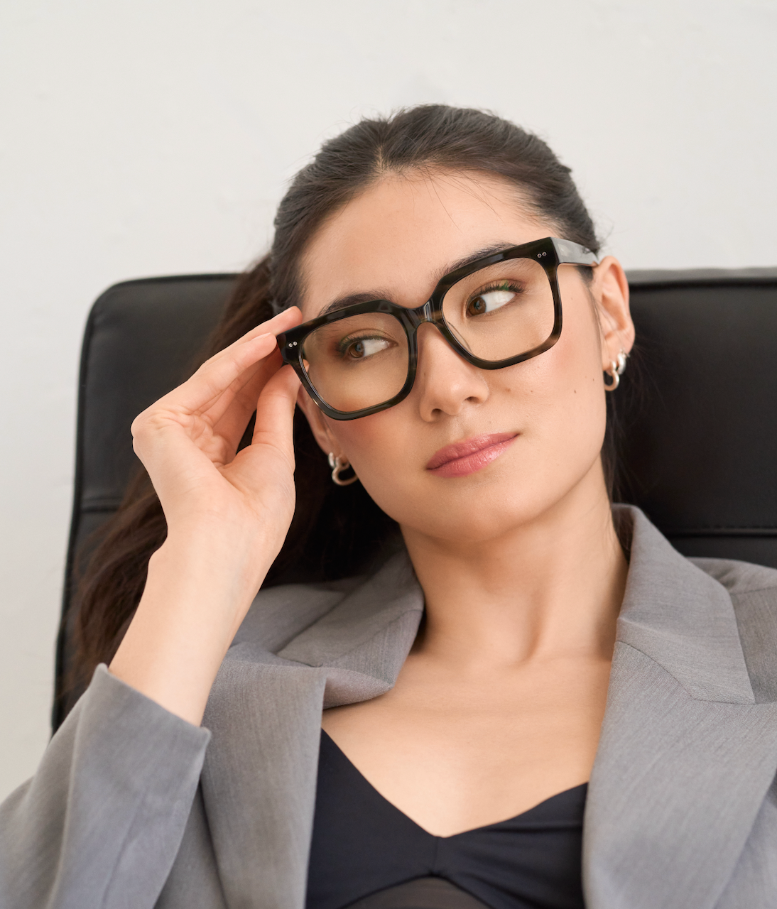 A woman wears KayTran Eyewears Andy - Graphite glasses and a gray blazer, seated with a neutral expression.
