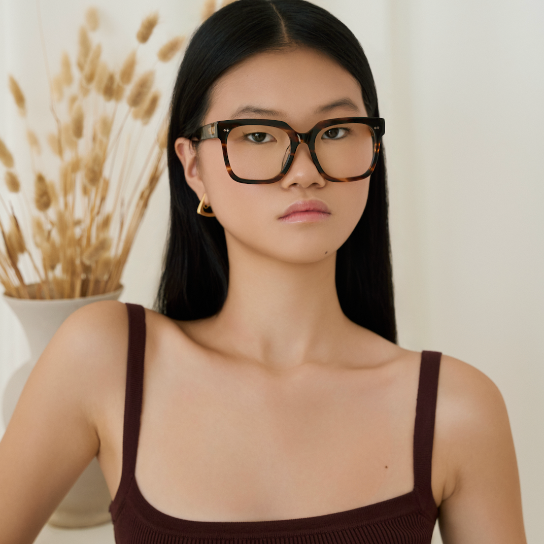 Woman with long black hair wears KayTran Eyewears Andy - Amber Honey glasses and a sleeveless top by a vase of dried grasses.