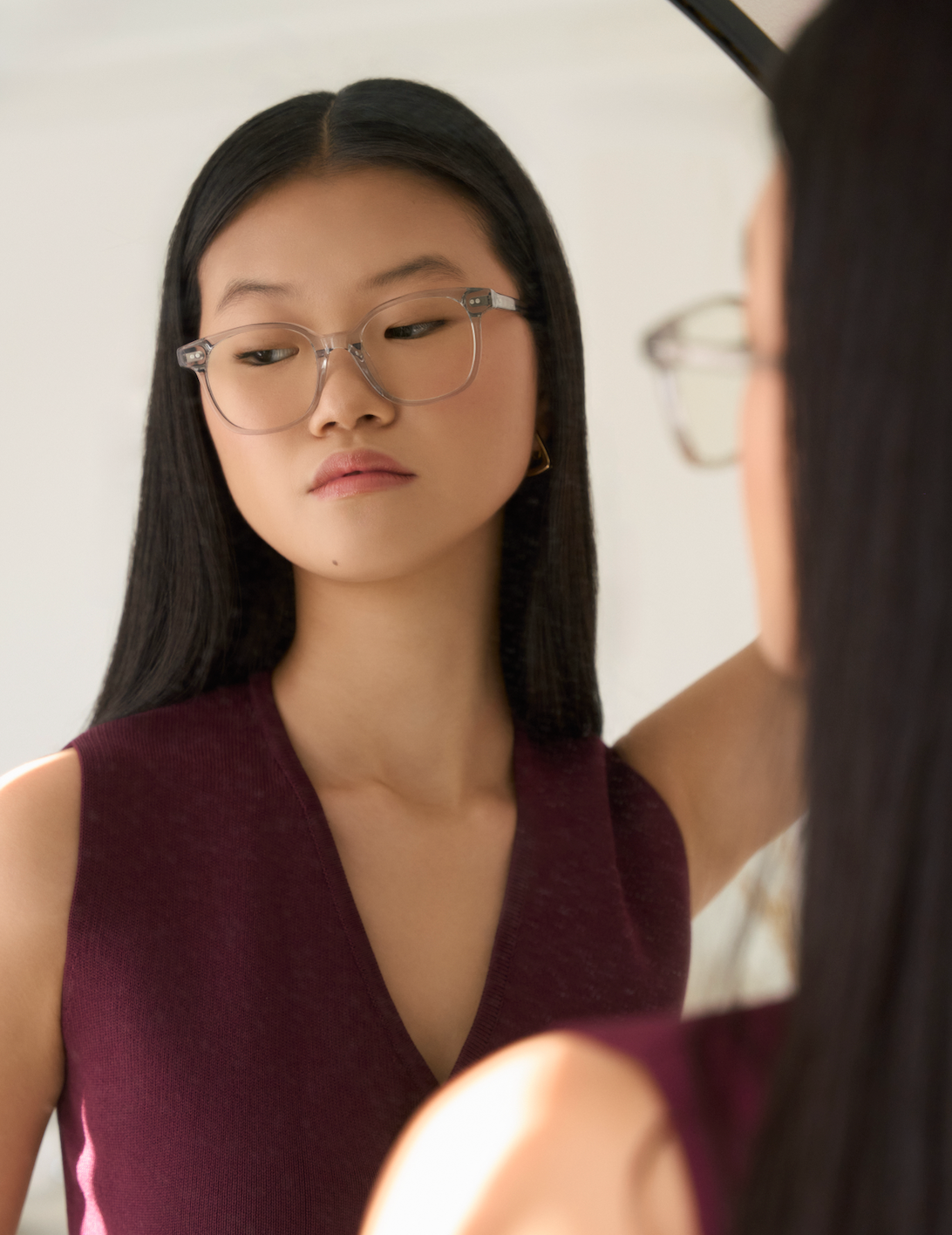 A woman with long straight hair wears KayTran Eyewear’s Easton - Cloud glasses and a sleeveless burgundy top in the mirror.