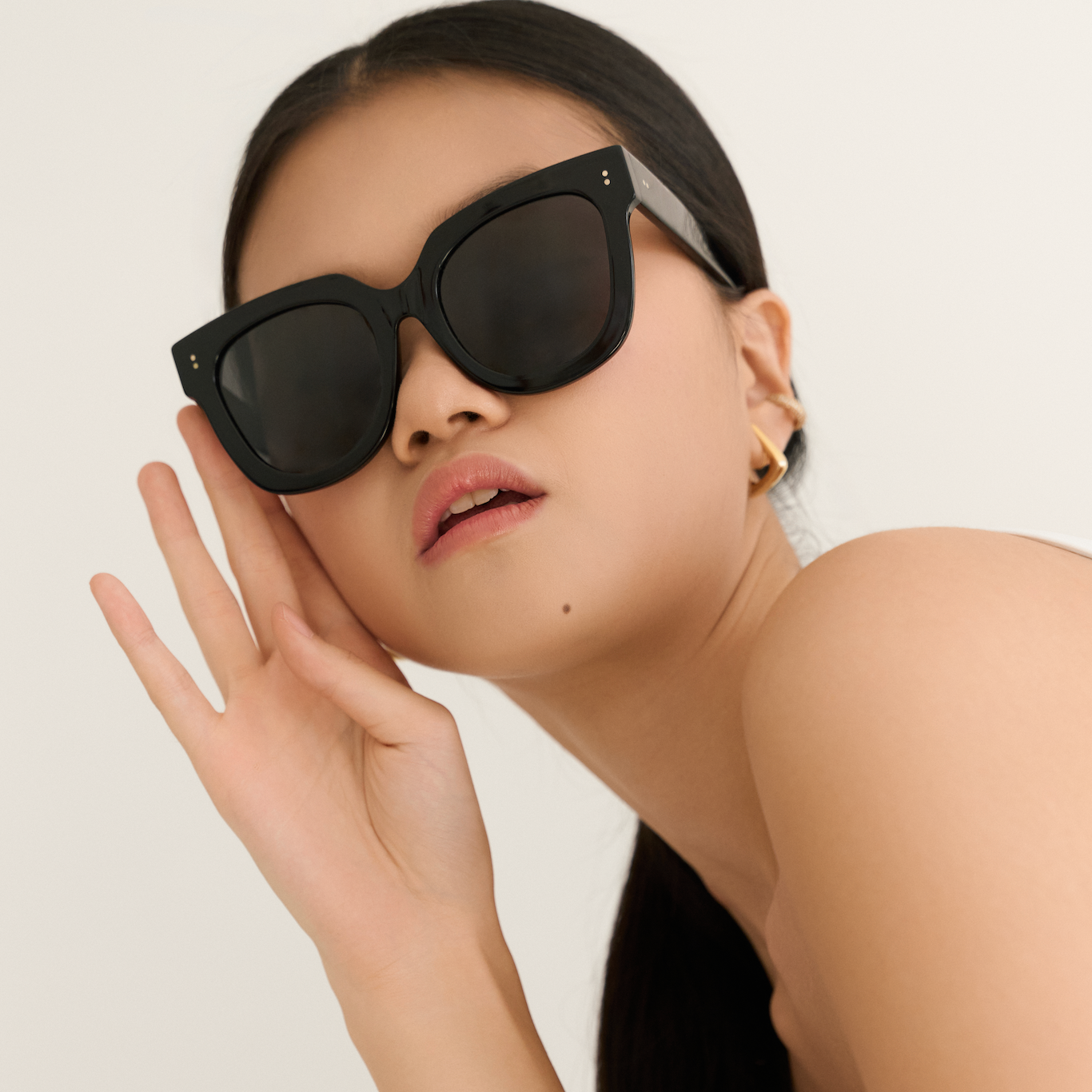 Woman wearing KayTran Eyewears Rowan - Polished Black sunglasses and gold earrings, posing against a plain background.