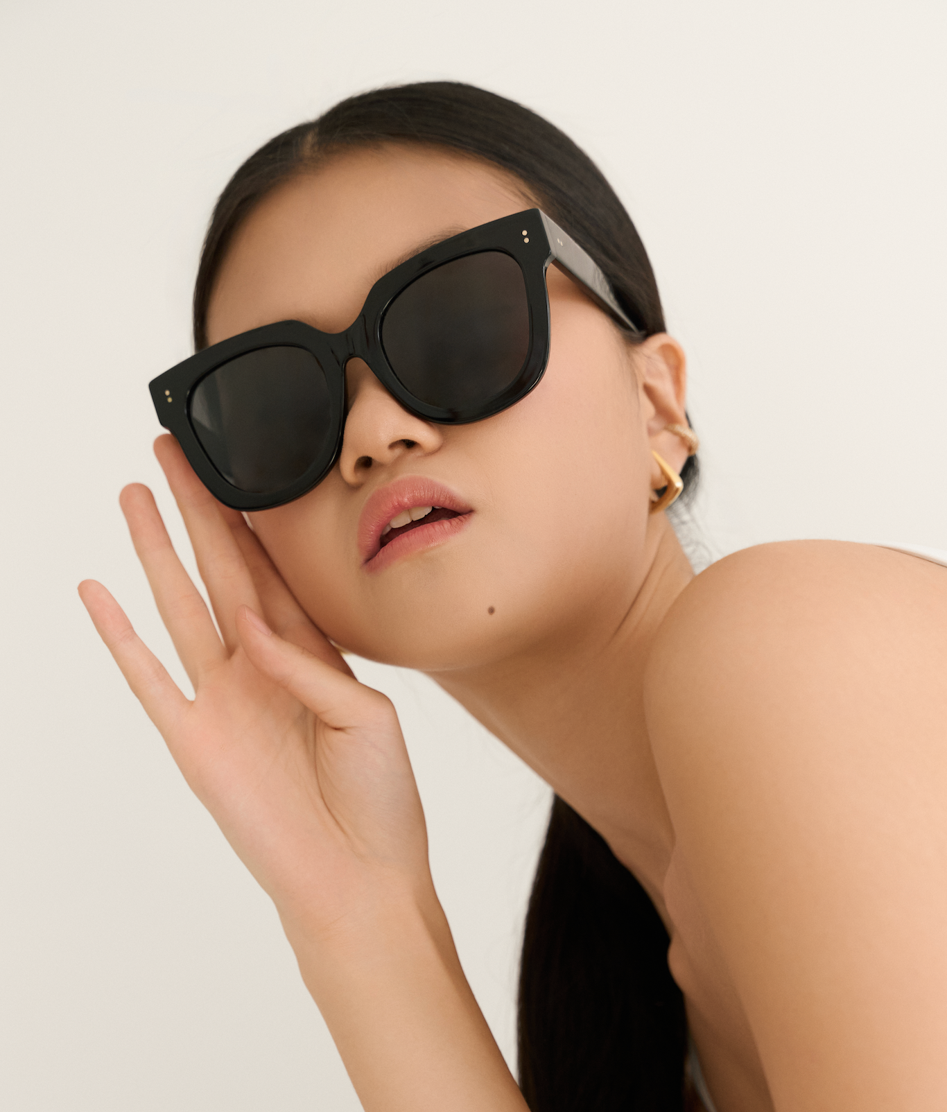Woman wearing KayTran Eyewears Rowan - Polished Black sunglasses and gold earrings, posing against a plain background.