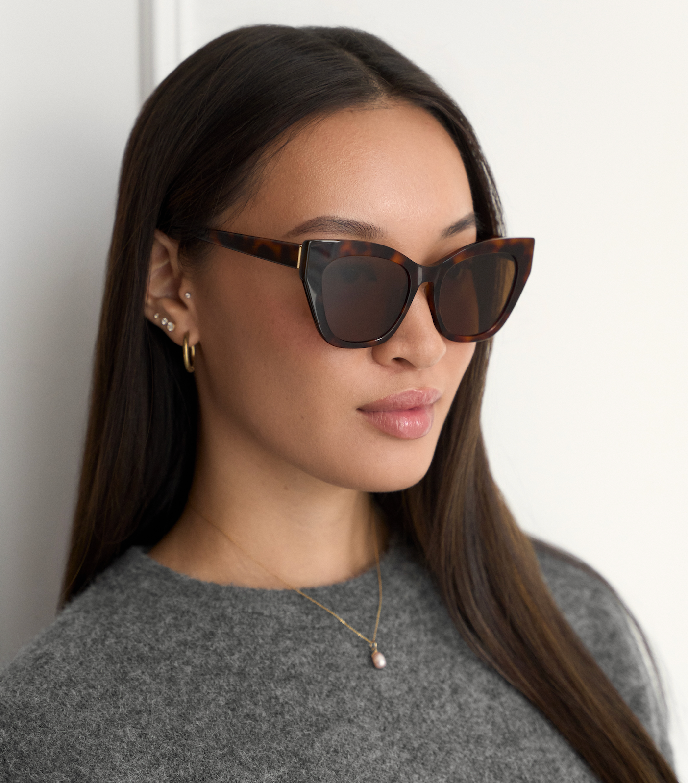 Woman with straight dark hair faces left wearing KayTran Eyewears Harper - Lava Tort cat-eye sunglasses and gold jewelry.