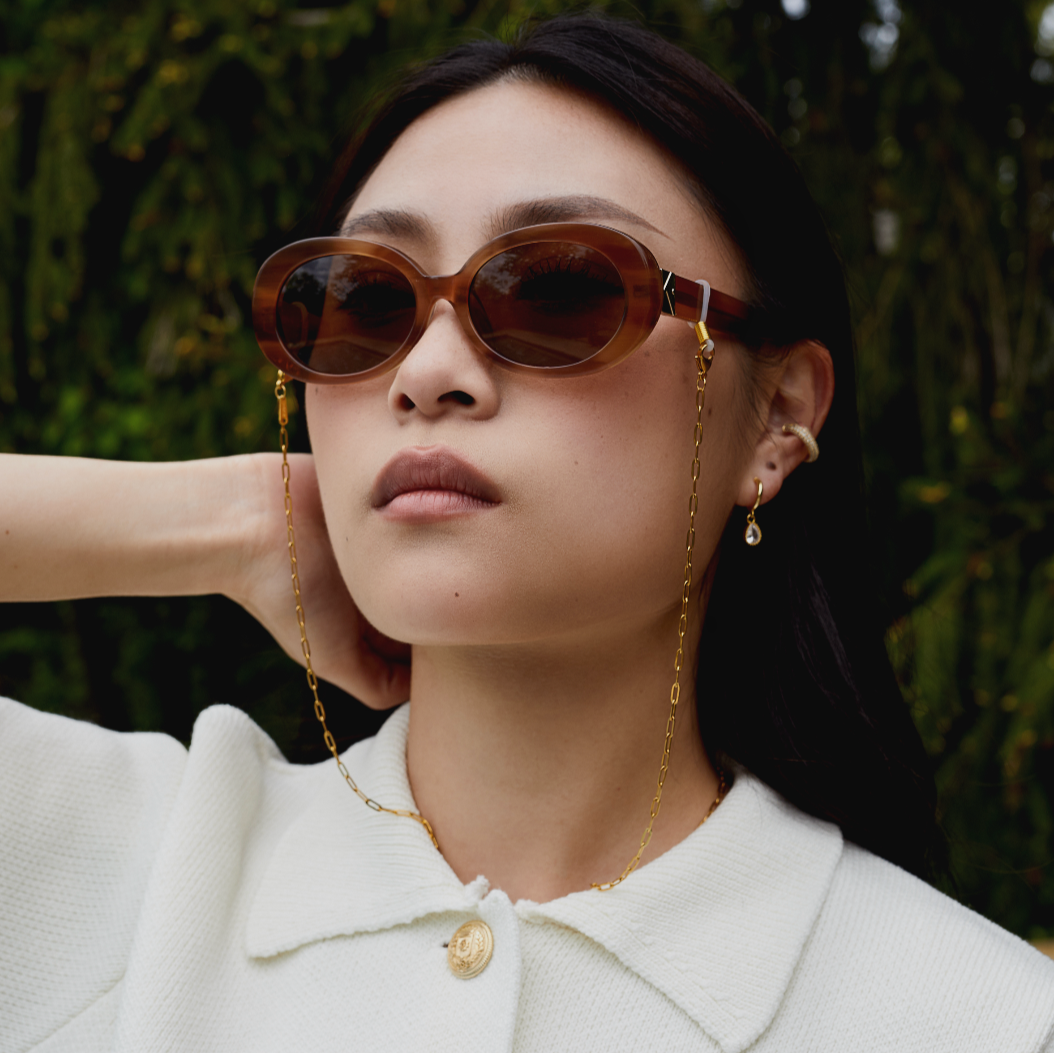 Woman in brown oval sunglasses, KayTran x Aiori Eyewear Chain, white collared top, gold earrings outdoors.