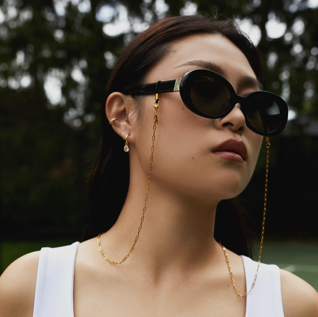 Woman wearing black KayTran Eyewear sunglasses with a gold KayTran x Aiori chain, white top, and small earring outdoors.