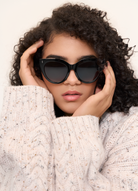 Woman with curly hair wears kaytran eyewears Aiden - Polished Black sunglasses and a cream knit sweater.