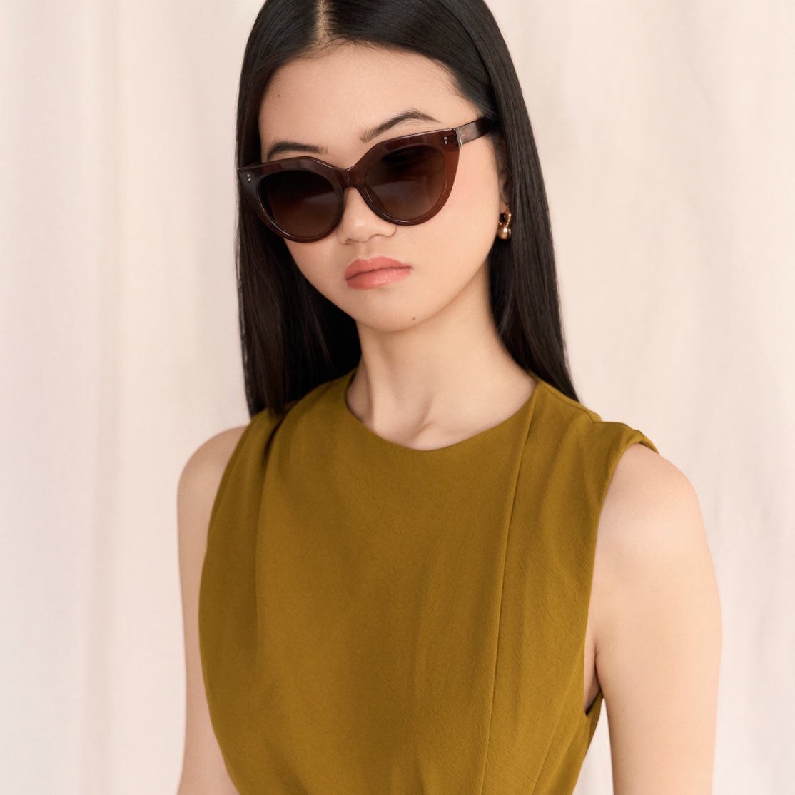 Woman with long black hair wears kaytran eyewear Aiden - Truffle sunglasses and a sleeveless olive dress.