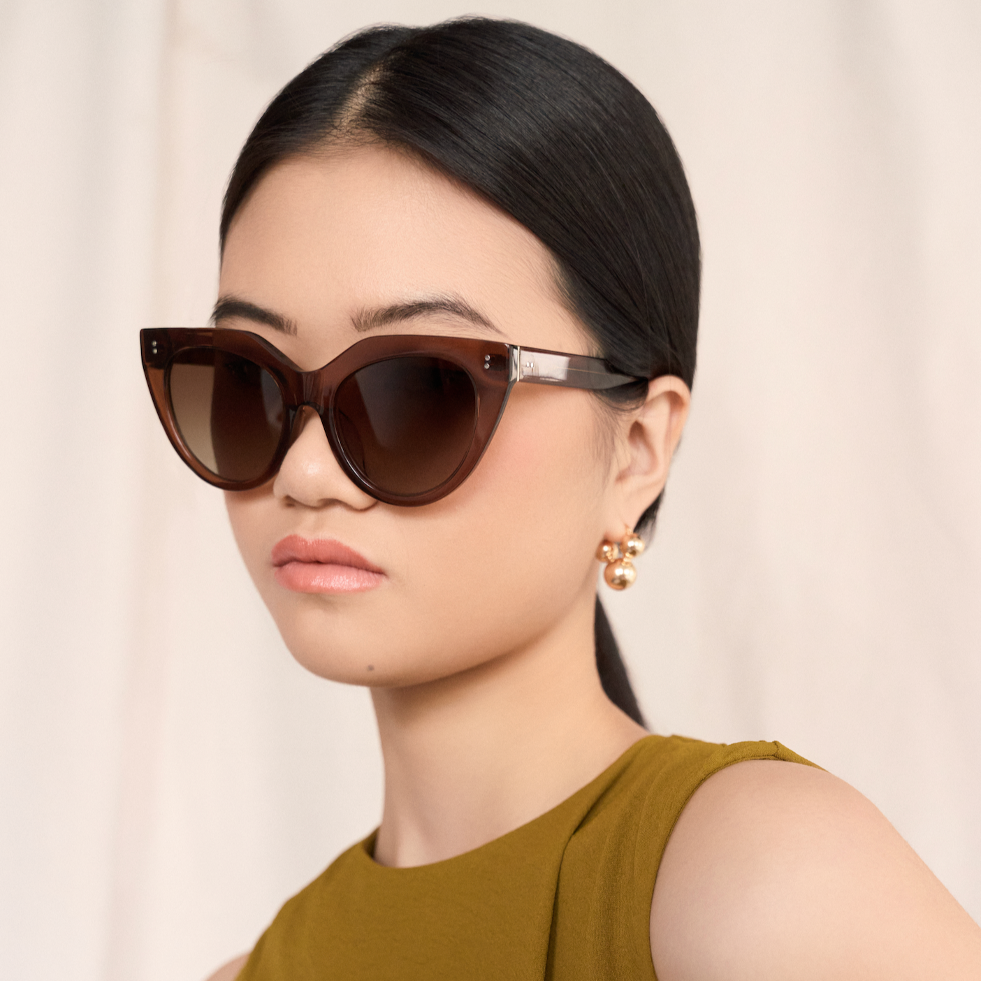 A woman with straight dark hair wears kaytran eyewears Aiden - Truffle sunglasses, gold earrings, and an olive top.