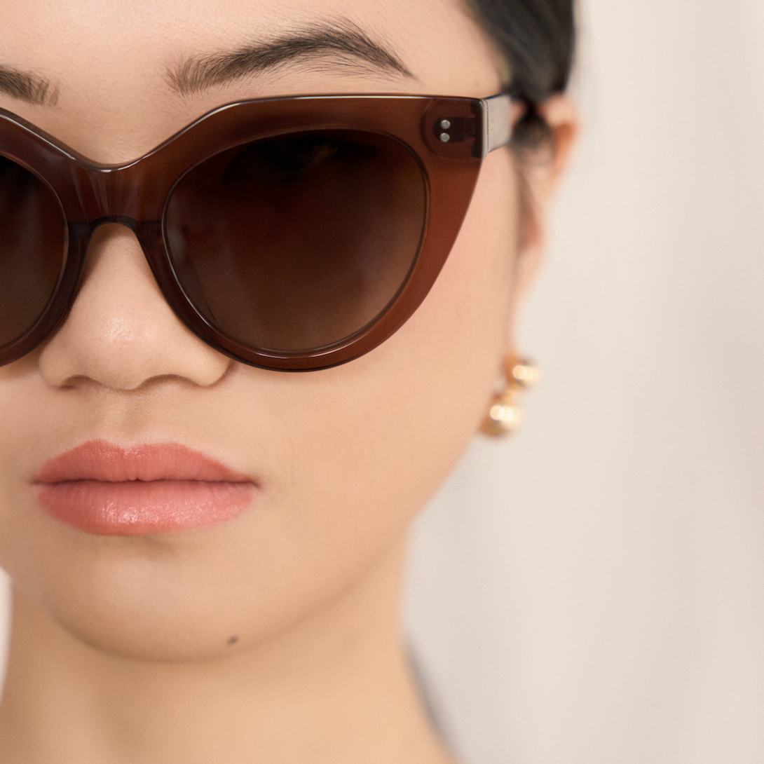 Close-up of a woman in kaytran eyewears Aiden - Truffle sunglasses, gold earrings, and a mustard yellow top.