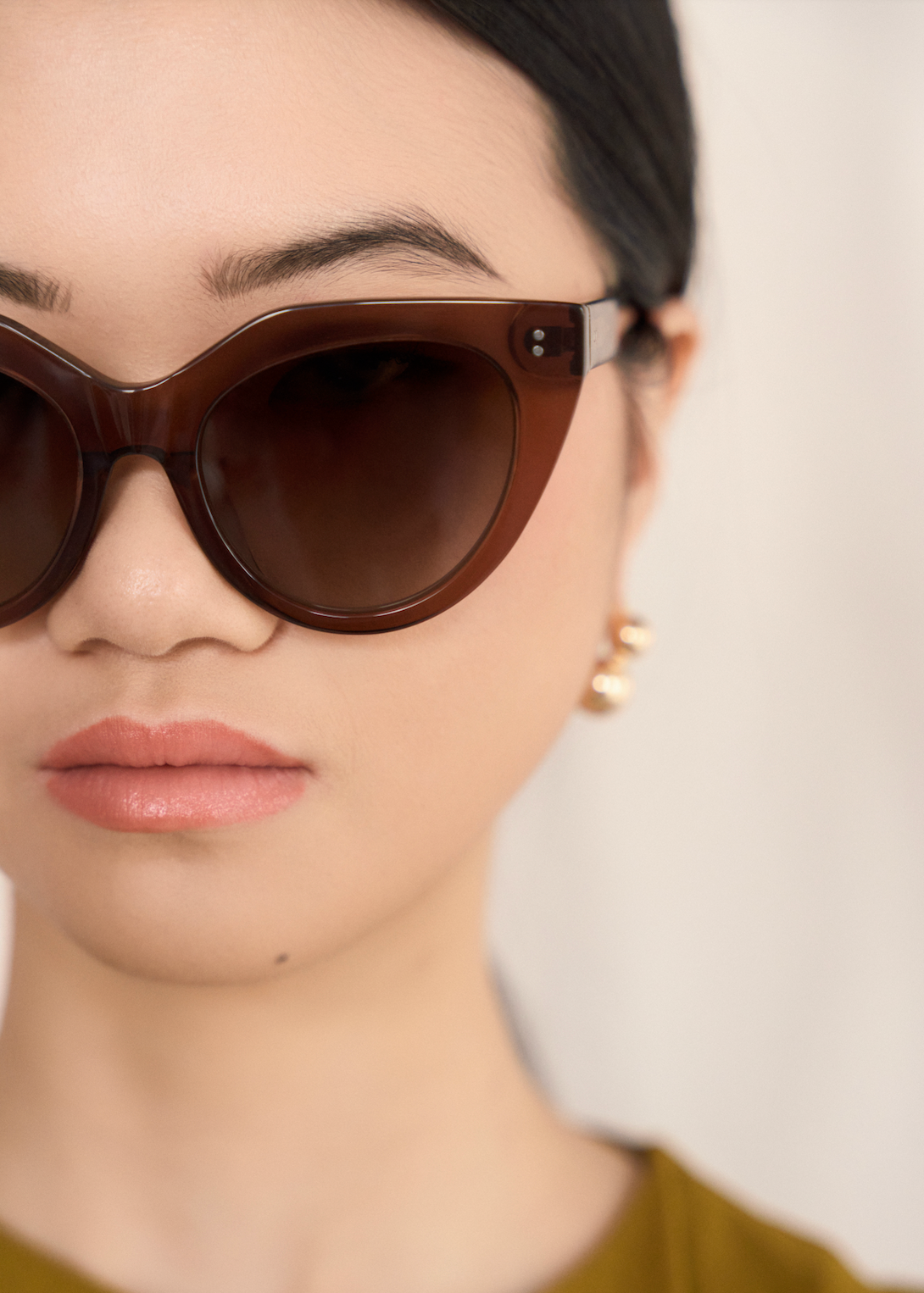 Close-up of a woman in kaytran eyewears Aiden - Truffle sunglasses, gold earrings, and a mustard yellow top.