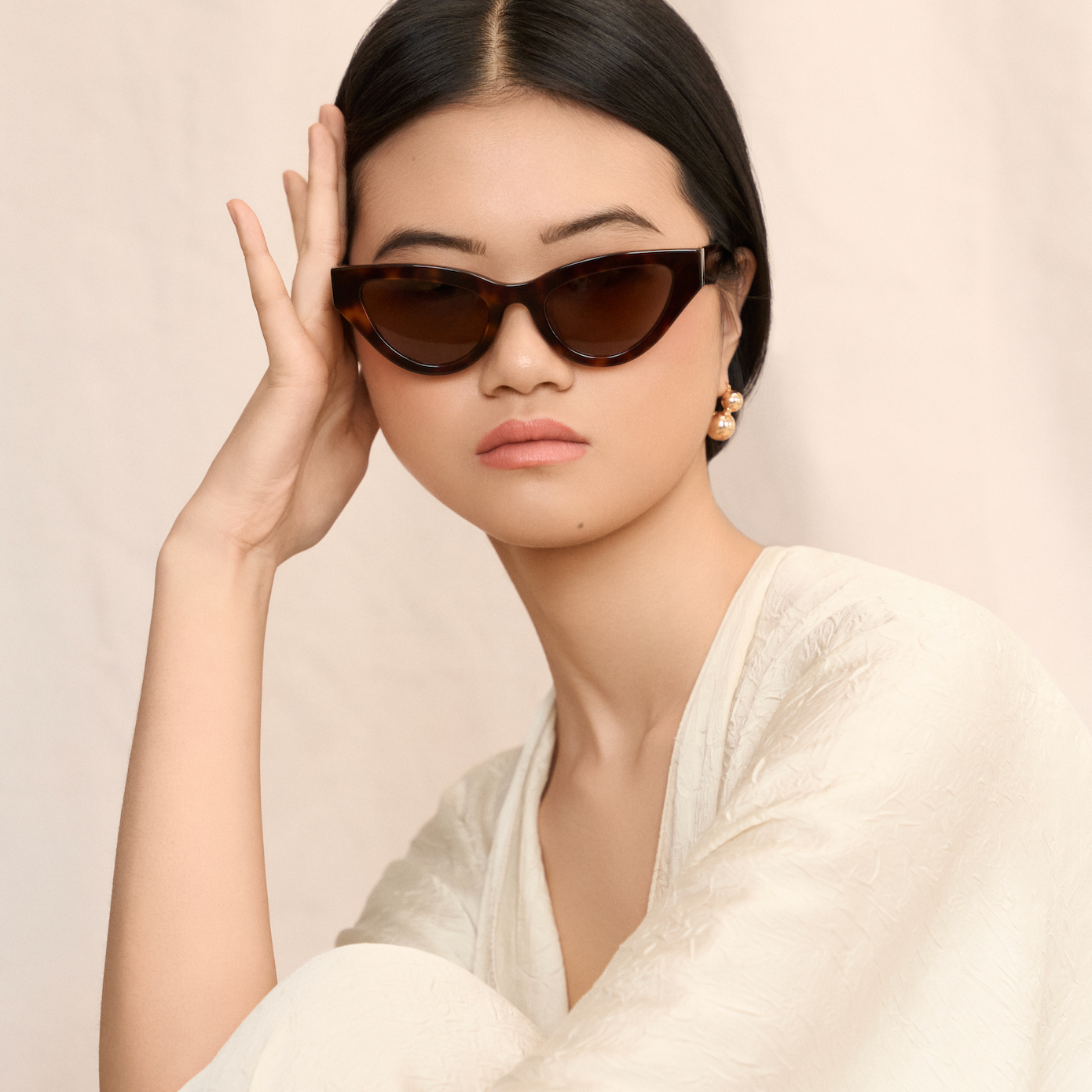 Woman in cream textured dress wears KayTran Eyewear Drew - Honey Tort cat-eye sunglasses, resting her head.