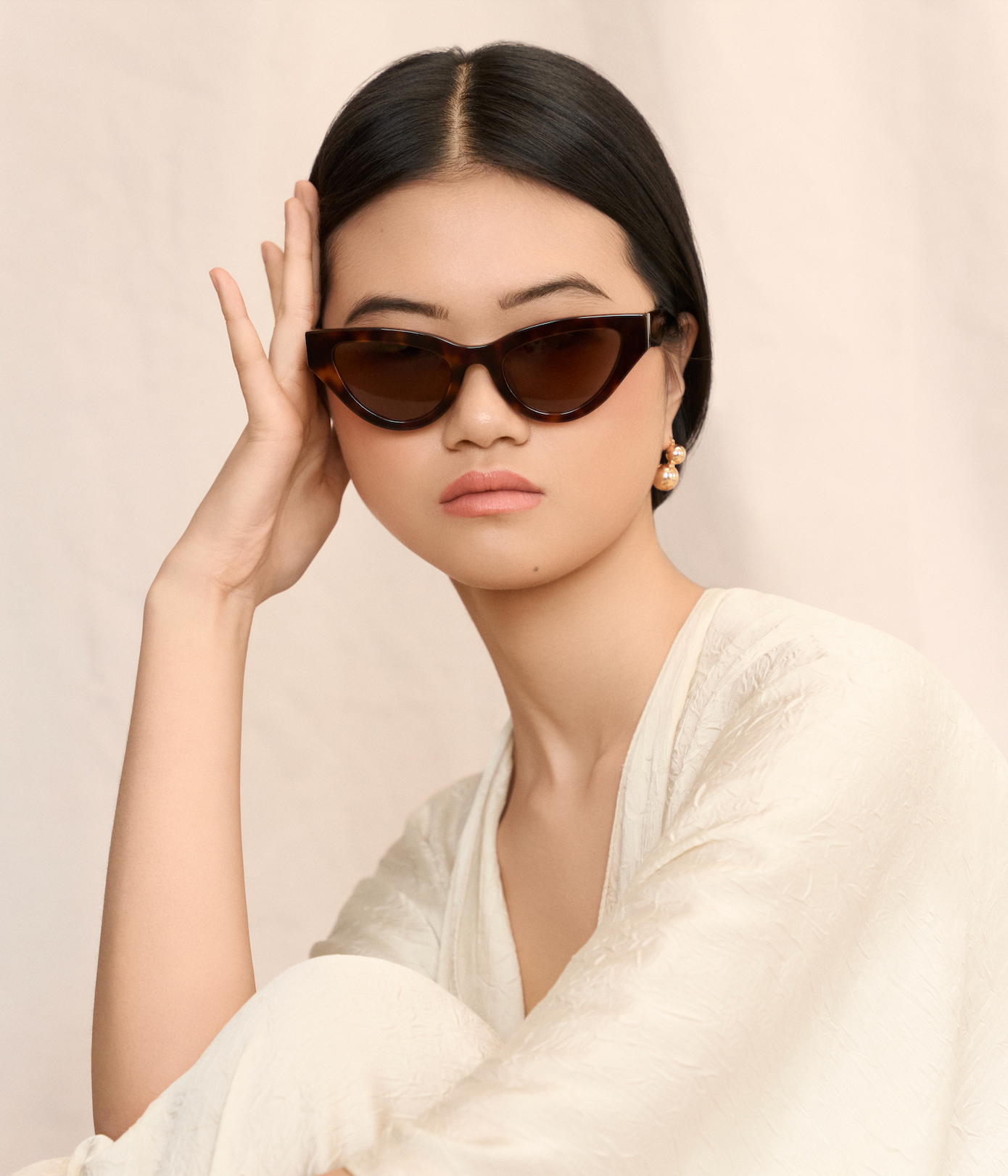 Woman in cream textured dress wears KayTran Eyewear Drew - Honey Tort cat-eye sunglasses, resting her head.
