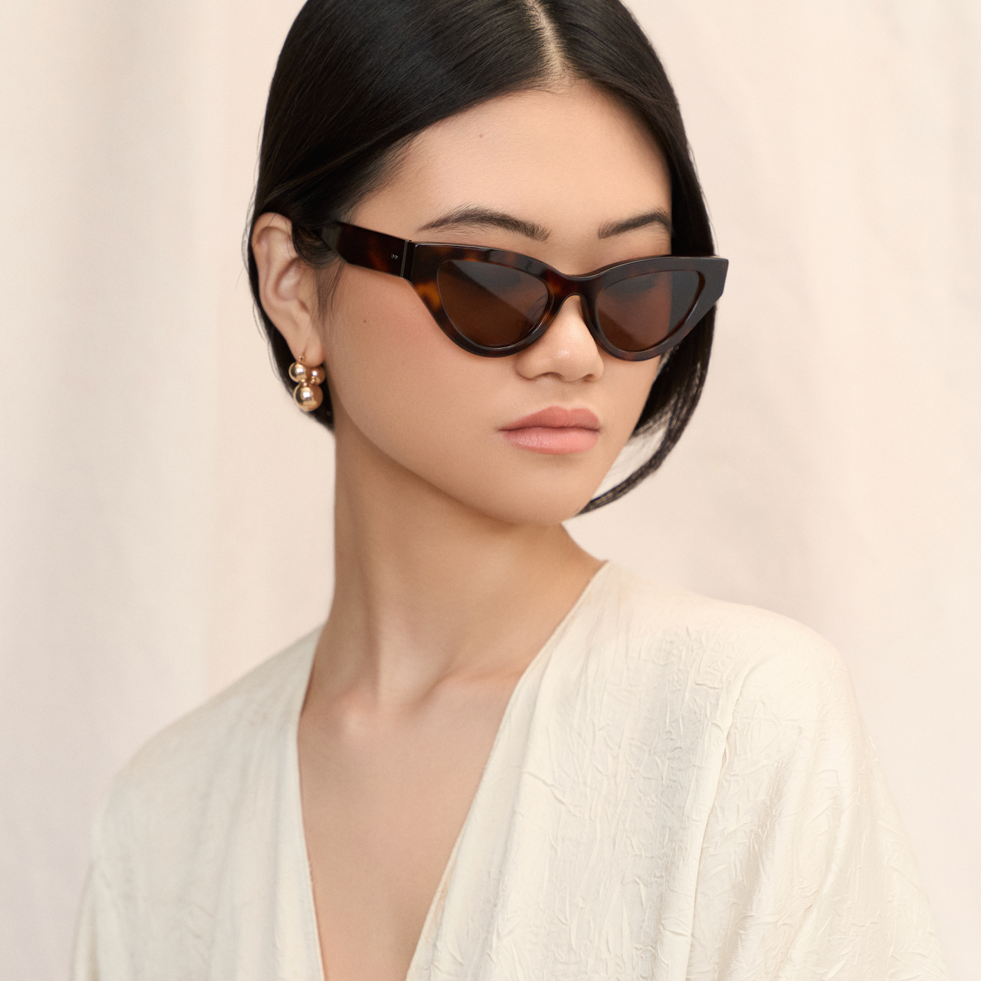 Woman in a cream top wears KayTran Eyewears Drew - Honey Tort cat-eye sunglasses, looking to the side.