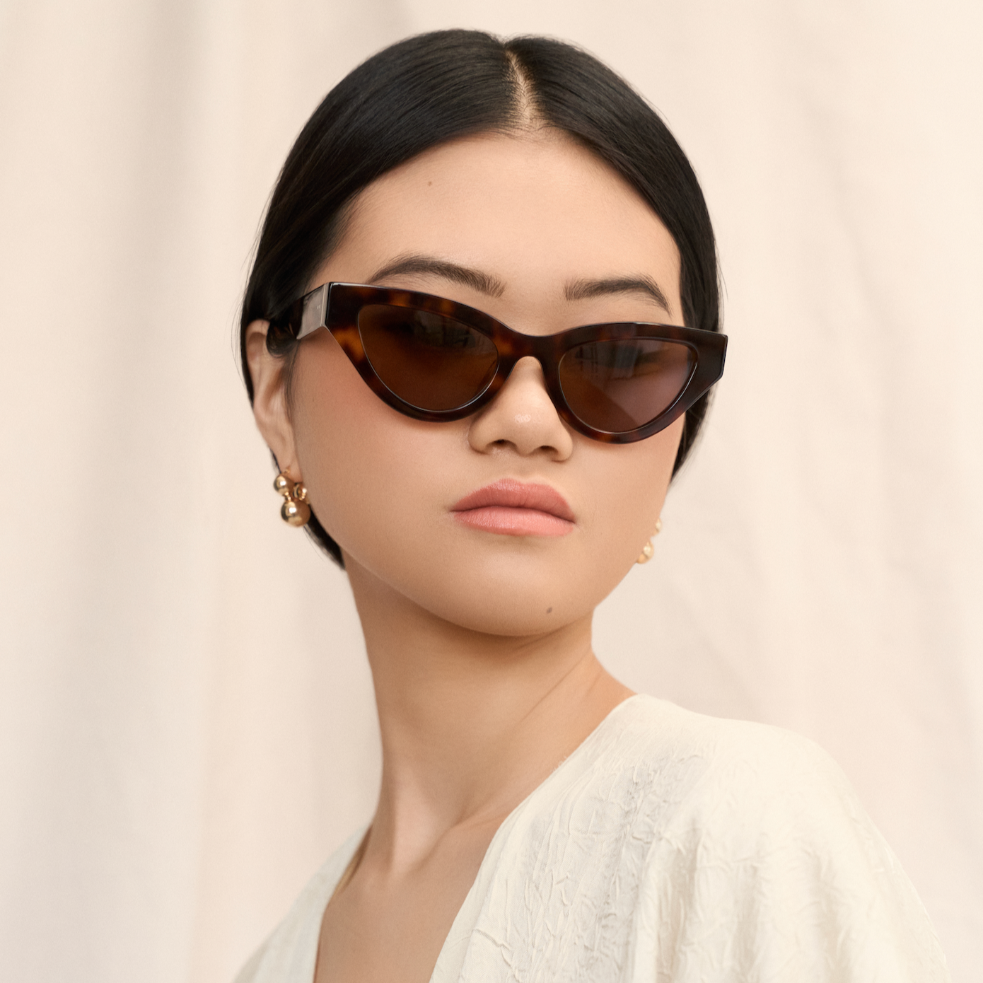 Woman with dark hair wears KayTran Eyewear Drew - Honey Tort cat-eye sunglasses and a cream textured top.