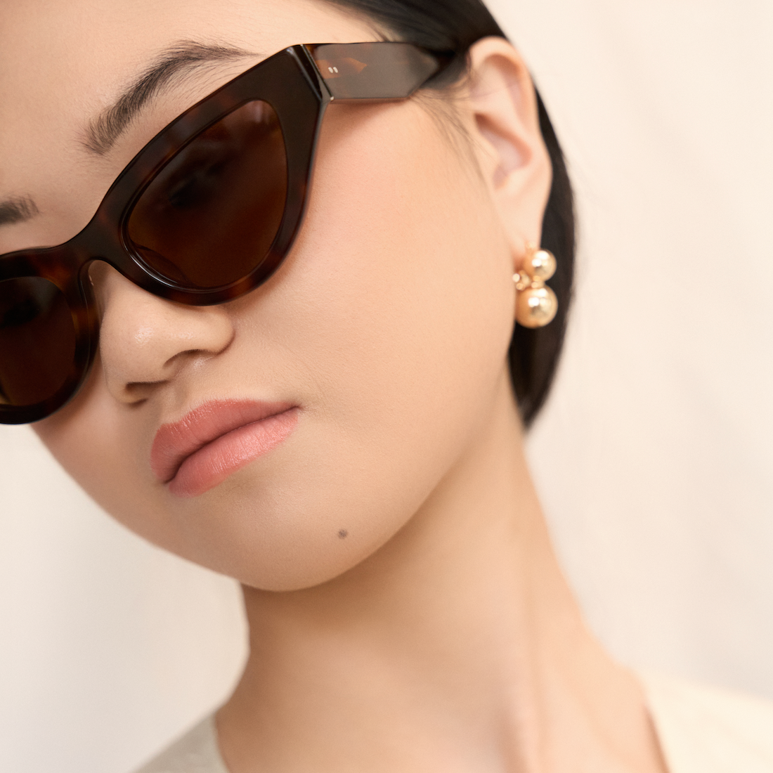 Woman with straight black hair wears KayTran Eyewear Drew - Honey Tort sunglasses and pearl earrings against a neutral backdrop.