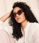 Woman with curly hair wearing KayTran Eyewear Drew - Burgundy sunglasses and a white blouse, resting her head on her hand.
