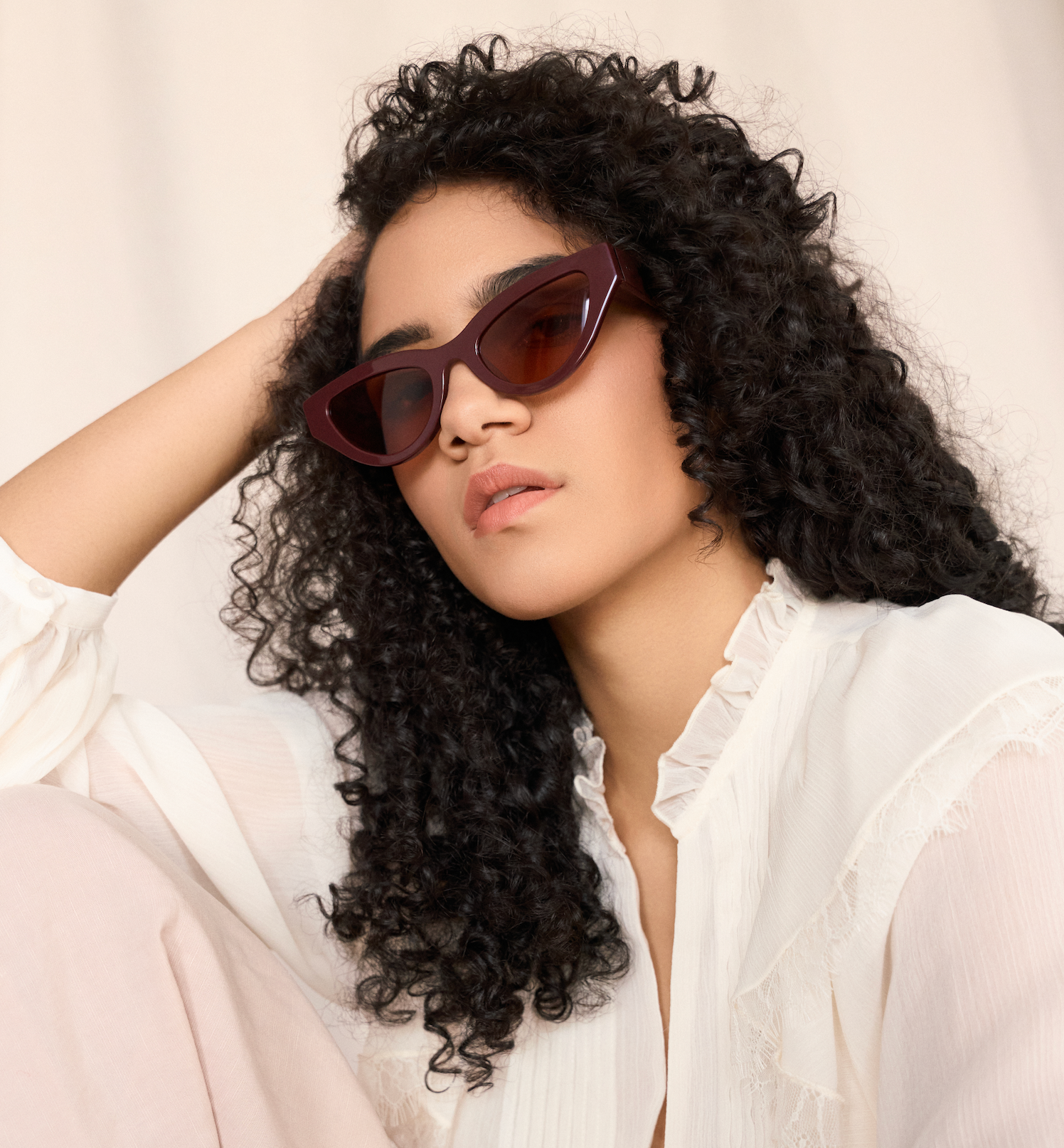 Woman with curly hair wearing KayTran Eyewear Drew - Burgundy sunglasses and a white blouse, resting her head on her hand.