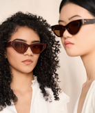 Two women in KayTran Eyewears Drew - Burgundy cat-eye sunglasses and white tops stand side by side against a neutral background.