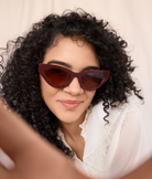 A woman with curly hair wears KayTran Eyewear’s Drew - Burgundy cat-eye sunglasses, smiling at the camera.