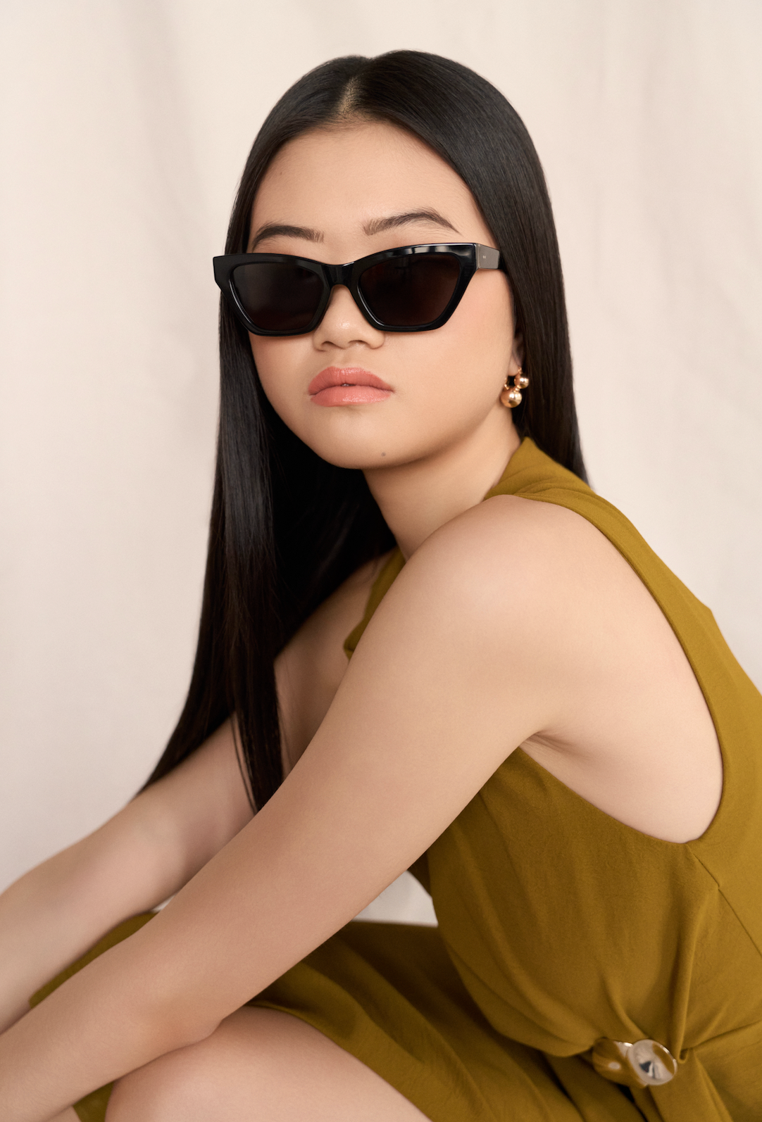 Woman with long black hair wears KayTran Eyewear Kai - Polished Black sunglasses and a sleeveless olive dress.