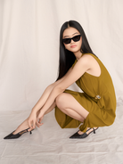 Woman in an olive green dress and black heels sits on the floor, wearing KayTran Eyewears Kai - Polished Black sunglasses.